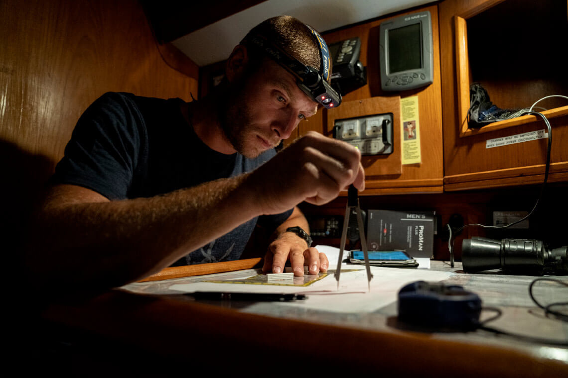 Natural Pharmaceuticals ProMan Plus Vyras burlaivio kajutėje naktį dirba su navigacijos žemėlapiu, naudodamas kompasą ir galvos žibintą. Šalia jo ant stalo matomi navigacijos prietaisai, žibintas ir dėžutė su „ProMan Plus“ maisto papildais.