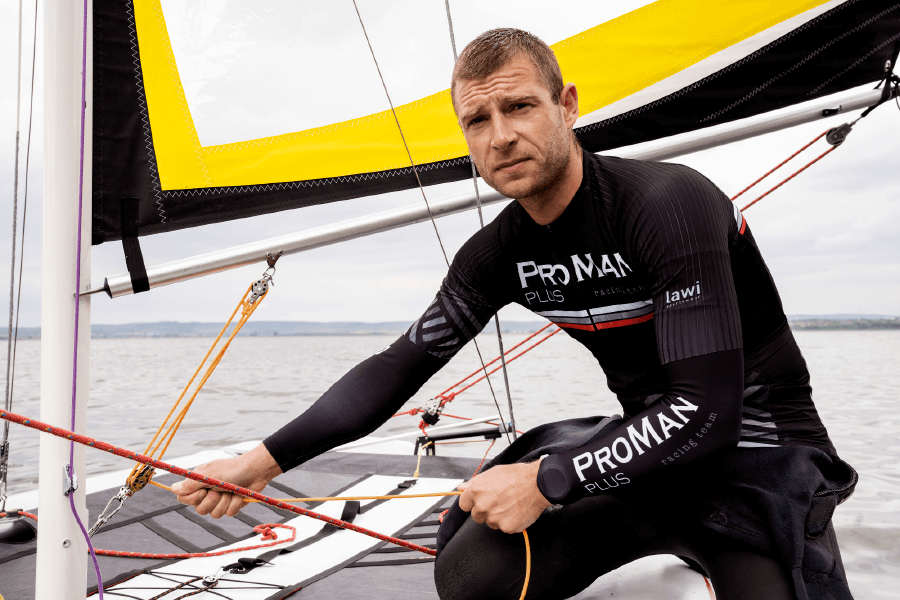 Natural Pharmaceuticals ProMan Plus Vyras su juodu nardymo kostiumu valdo burlaivio stiebą ir virves ramioje ežero aplinkoje.