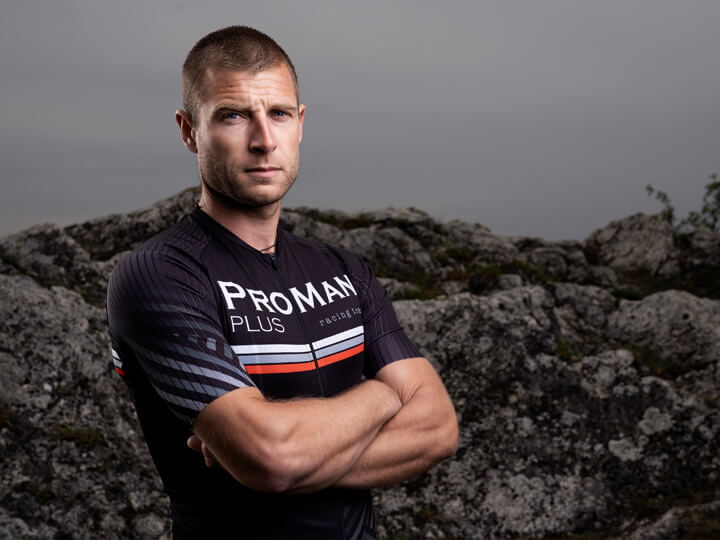 Natural Pharmaceuticals ProMan Plus Vyriškis dėvi „ProMan Plus“ dviračių aprangą, stovėdamas susikryžiavęs rankas prieš rūsčią uolėtą foną.