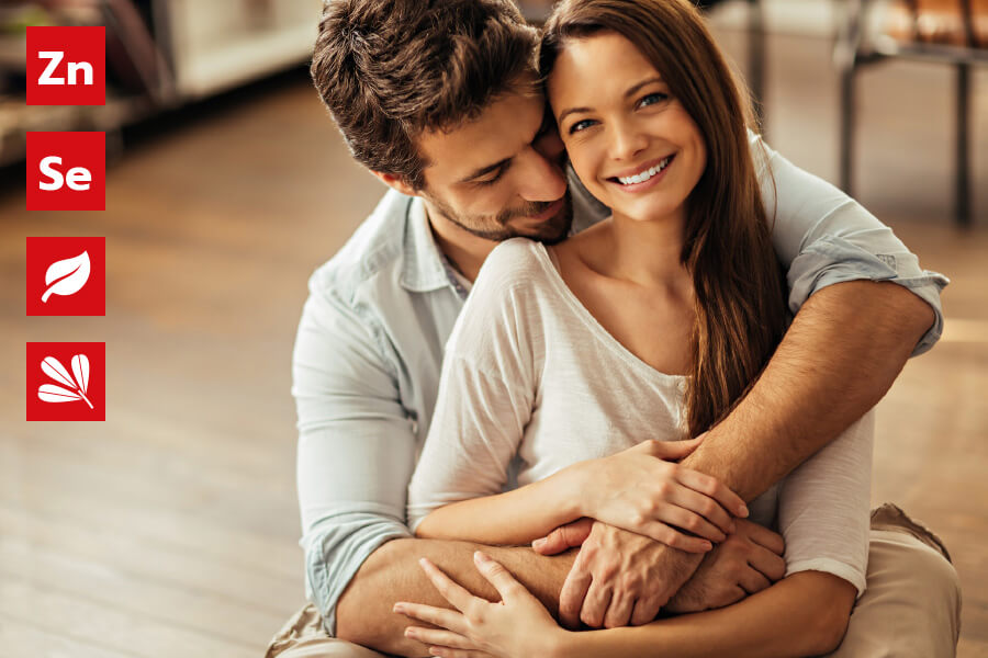 Natural Pharmaceuticals ProMan Plus Jaunas vyras ir moteris sėdi kartu, apsikabinę ir šypsodamiesi šiltoje, jaukioje aplinkoje. Kairėje pusėje yra raudonos piktogramos, simbolizuojančios ingredientus: Zn (cinkas), Se (selenas) ir lapų simbolis, reiškiantis augalų ekstraktus.