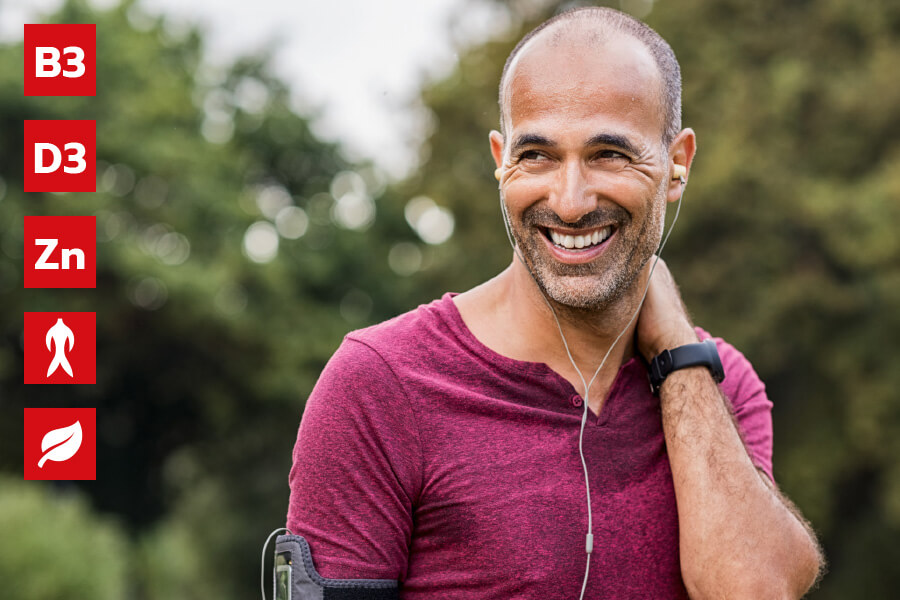 Natural Pharmaceuticals ProMan Plus Šypsantis vidutinio amžiaus vyras, vilkintis sportinį marškinėlį, ausines ir apyrankę, stovi lauke, apsuptas žalumos. Kairėje yra raudonos piktogramos ir simboliai, žymintys ingredientus: vitaminą B3, vitaminą D3, cinką (Zn), ženšenį ir augalų ekstraktus.